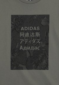 Harmaa t-paita, jossa on musta graafinen kuva, jossa on teksturoituja lehtiä ja sana "ADIDAS" useilla kielillä. Pyöreä kaula-aukko, pehmeä kangas.