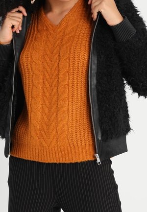 Vrouw draagt een oranje kabelgebreide trui onder een deels dichtgeritste zwarte textuurjas en zwarte pantalon met krijtstreep.