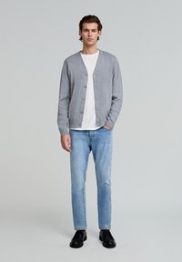 Grå cardigan med knapper, båret over en hvid t-shirt, parret med lyseblå jeans og sorte sko. Ensfarvet baggrund.