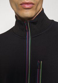 Veste zippée noire avec col haut, dotée de zippers multicolores et d'une petite poche zippée sur la poitrine. Texture lisse, design ajusté.