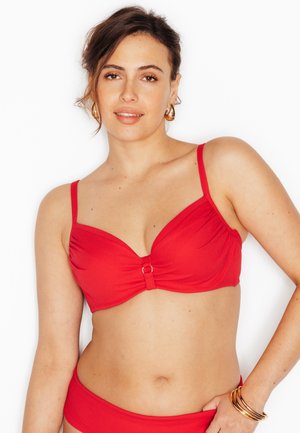 Femme à la peau claire et aux cheveux bruns portant un haut et un bas de bikini rouges, des boucles d'oreilles créoles dorées et plusieurs bracelets dorés au poignet droit.