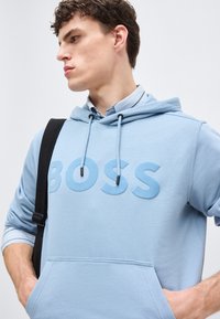 Jeune homme portant un sweat à capuche bleu clair avec l'inscription "BOSS", superposé à une chemise à col, portant une sangle noire sur une épaule.