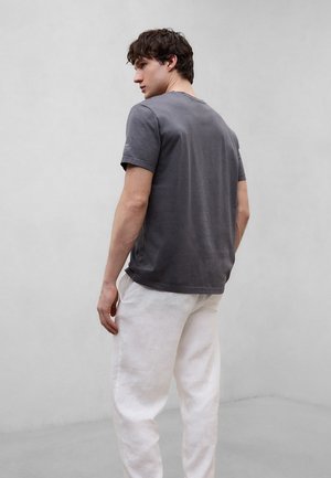 Junger Mann, der nach links blickt, trägt ein dunkelgraues kurzärmliges Hemd und lockere weiße Hose, steht vor einem schlichten hellgrauen Hintergrund.