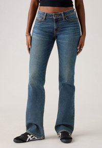 Femme portant un jean bootcut bleu, un haut court noir et des baskets noires à rayures blanches, debout les mains le long du corps.