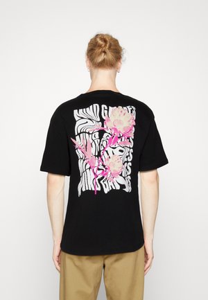 Person with blonde hair in a bun wearing a black t-shirt with white wavy text and pink floral design on the back, standing against a plain white background.