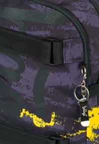 Zaino viola con motivi grafici in grigio scuro e accenti gialli. Presenta spallacci neri, un tirante in metallo argentato e un tessuto testurizzato.