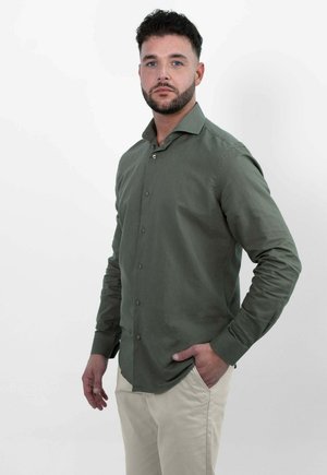 Man met kort krullend donker haar en baard, draagt een olijfgroen overhemd met knopen en lichte beige broek, staat met één hand in de zak.