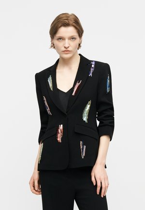 Nő fekete blézerben, amely színes flitterekkel van díszítve, fekete nadrággal, egyszínű világos háttér előtt állva.