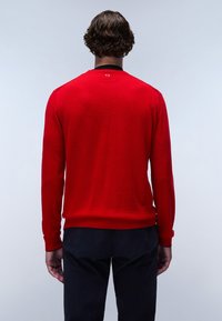 Suéter de lana rojo con mangas largas y puños acanalados, que presenta un escote negro y un sutil detalle del logotipo en la parte posterior. Combinado con pantalones oscuros.