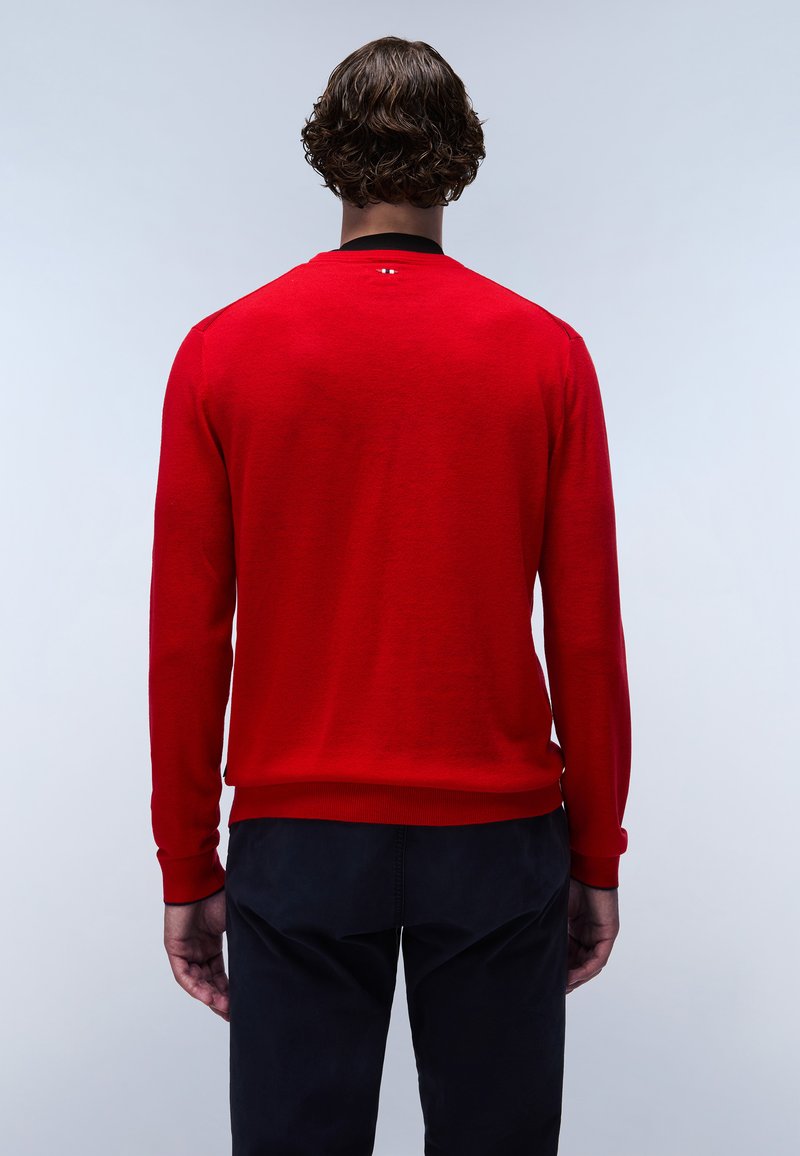 Czerwony sweter dzianinowy z długimi rękawami i ściągaczami, z czarną dekoltem i subtelnym logotypem z tyłu. Połączony z ciemnymi spodniami.