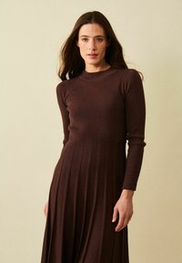Ruskea ribattu pitkähihaine mekko, jossa on tiukka yläosa ja laskostettu hame. Sujuva tekstuuri, suora siluetti ja pyöreä pääntie.