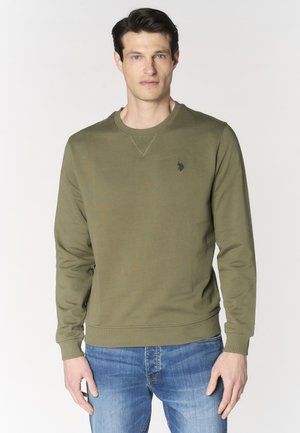 Uomo che indossa una felpa verde oliva a maniche lunghe e jeans blu, in piedi davanti a uno sfondo chiaro e semplice.