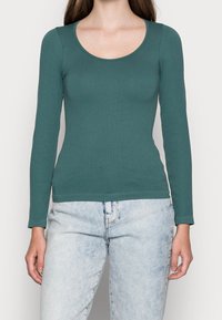 Langärmeliges, geripptes Oberteil in Türa, mit Rundhalsausschnitt. Aus einem weichen, dehnbaren Stoff gefertigt, kombiniert mit hellblauen, verwaschenen Jeans.