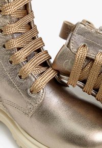 Metallicgoldene Knöchelstiefel mit dicken, geflochtenen Schnürsenkeln, strukturiertem Leder und einer klobigen weißen Sohle. Mit Metallösen und Branding-Element am Absatz.