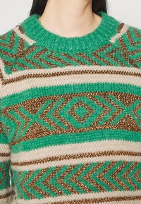 Pull en maille épaisse avec des motifs géométriques verts, beiges et bronze métallisé et un col côtelé vert, porté par une personne.