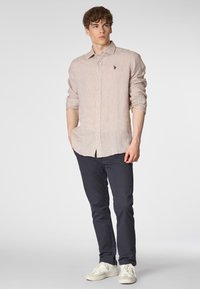 Camicia a maniche lunghe beige chiaro con colletto button-down, abbinata a pantaloni blu navy e scarpe bianche. Presenta un piccolo logo ricamato.