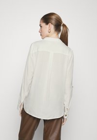 Light beige blouse with a smooth fabric, long sleeves, and a collar. Features a curved hem and button accents on the sleeves.