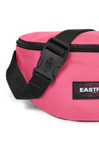 Vaateva pinkki vyölaukku mustalla säädettävällä hihnalla, jossa on vetoketjulukko ja logotikki mustalla taustalla. Kuosis fabric.