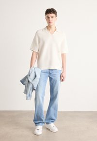 Strikket kremfarget kortermet polo-skjorte med V-hals, kombinert med lyseblå jeans og holder en falmet denimjakke. Hvite joggesko fullfører antrekket.