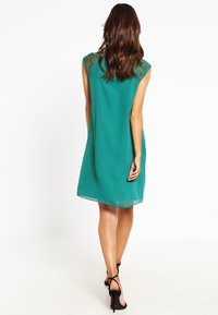 Groene mouwloze jurk met kantaccenten op de schouders, vloeiende stof, knie lengte ontwerp en een licht uitlopende silhouet. Zwarte sandalen met bandjes.