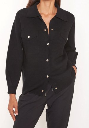 Femme portant une chemise noire boutonnée avec poches poitrine et manches longues, associée à un pantalon noir, posant avec une main dans la poche.