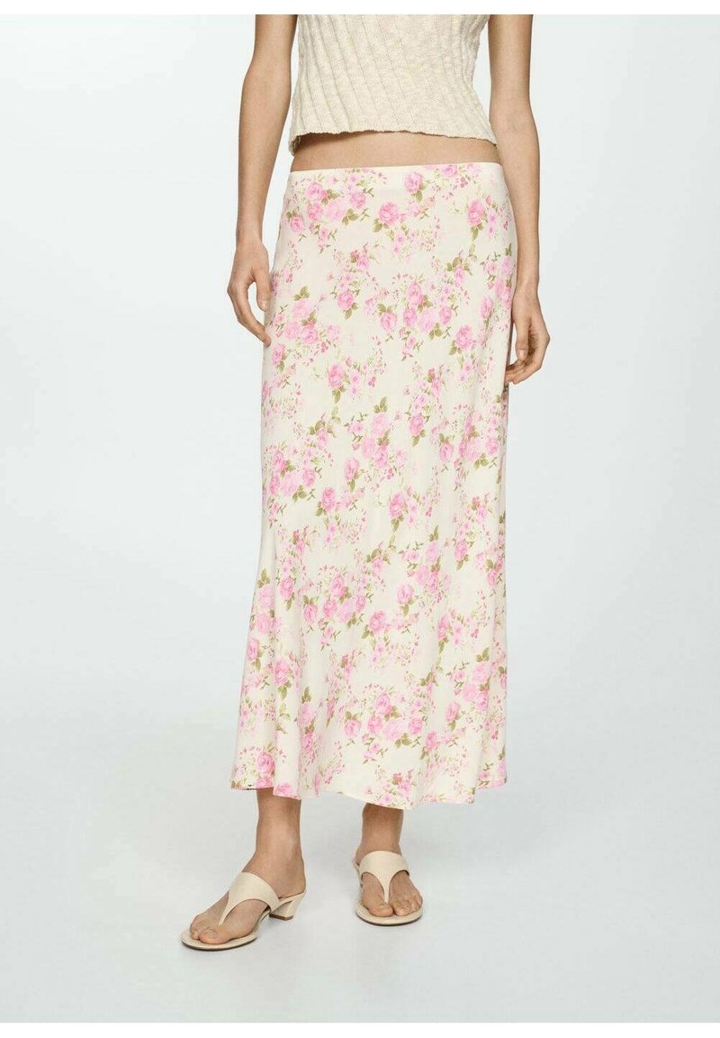 Femme portant une jupe longue crème à fleurs roses, des sandales à talons beiges et un top court sans manches sur fond uni.