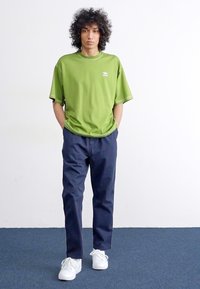 T-shirt vert surdimensionné à manches courtes, avec un petit logo, associé à un pantalon bleu marine et des baskets blanches. Fond neutre.