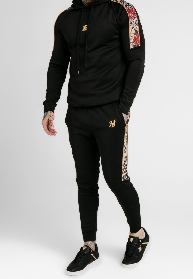 Mann trägt schwarzen Trainingsanzug mit floralen und Leopardenmuster-Streifen an Ärmeln und Hosenbeinen, kombiniert mit schwarzen Sneakers.