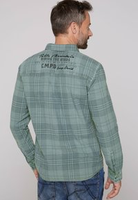 Camisa de cuadros verdes con mangas largas, cuello abotonado y texto impreso en la parte posterior; presenta costuras mínimas y un dobladillo curvado.