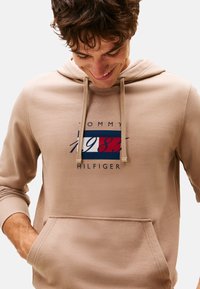 Joven mirando hacia abajo, sonriendo, con las manos en los bolsillos, vistiendo una sudadera beige de Tommy Hilfiger con el logo de 1985 en el frente.