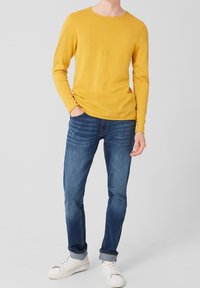 Homme portant une chemise jaune moutarde à manches longues, un jean bleu retroussé, des baskets blanches, debout avec une main dans la poche sur un fond uni.