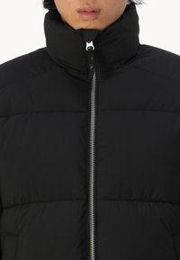 Osoba ubrana w czarną pikowaną kurtkę z wysokim kołnierzem zapinanym na zamek, widoczne tylko dolna część twarzy i szyja.