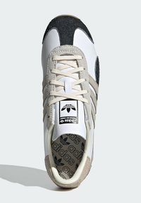 Sneaker bianco con accenti in suede grigio e nero, punta rotonda, lacci crema e logo in rilievo sulla linguetta. Presenta suola in gomma e interno pattern.