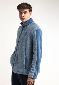 Chaqueta de forro polar azul con un cuerpo texturizado y mangas lisas, que presenta un cuello alto, cierre de cremallera y bolsillos laterales.