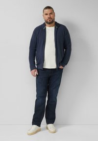 Marineblauw jack met rits over een wit T-shirt, gecombineerd met donkere spijkerbroek en witte sneakers. Gladde textuur, losse pasvorm, zonder zichtbare patronen.