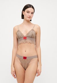 Béžový krajkový bralet a odpovídající tanga s vlnkovými okraji. Bralet má červený pás s logem 'HUGO'. Materiál vypadá jemně a strukturovaně.