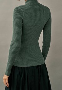 Pull à col roulé côtelé de couleur vert foncé, cintré à la taille, manches longues, associé à une jupe noire, vu de dos.