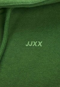 Primer plano de una tela verde con un cordón trenzado verde y texto "JJXX" en verde claro impreso cerca del centro superior.