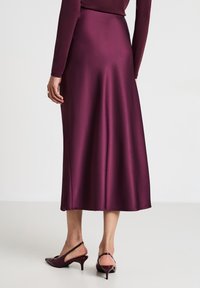 Gonna di satin bordeaux a metà lunghezza con una texture liscia. Presenta una forma svasata e uno spacco laterale. Abbinata a tacchi a punta scura.