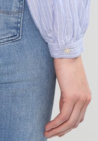 Helle blaue gestreifte Hemdmanschette mit gerafftem Stoff und einem Knopfdetail, kombiniert mit hellblauen Denim-Jeans. Weiche Textur und strukturiertes Design.