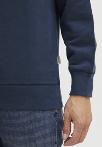 Felpa blu navy con polsini e orlo a coste. Presenta un piccolo marchio sul lato. Lo sfondo è semplice e neutro.