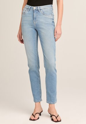 Frau trägt hellblaue Jeans mit geradem Bein und schwarze offene Sandalen mit Absatz, steht vor neutralem Hintergrund.