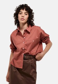 Chemise boutonnée couleur rouille avec col, poche poitrine unique, manches remontées et texture lisse. Associée à un pantalon marron taille haute.