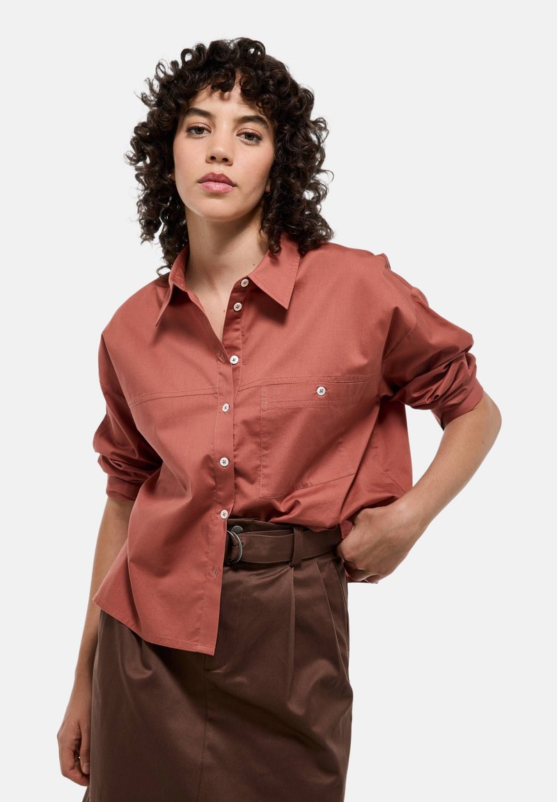 Chemise boutonnée couleur rouille avec col, poche poitrine unique, manches remontées et texture lisse. Associée à un pantalon marron taille haute.