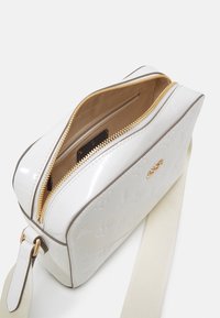 Bílá lakovaná crossbody taška se zlatým zipem, částečně otevřená, aby odhalila béžový interiér. Obsahuje embosovaný vzor a nastavitelný krémový popruh.