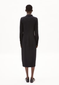 Schwarzes, langärmliges Kleid mit Kragen, schmal geschnitten, knielang, glattes Material, subtile Nahtdetails, kombiniert mit schwarzen Schuhen.