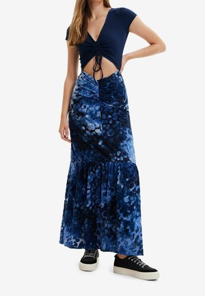 Vrouw draagt een marineblauw geplooid cropped top met een knoop aan de voorkant en een lange blauwe rok met een patroon, gecombineerd met sneakers, staand met één hand op de heup.