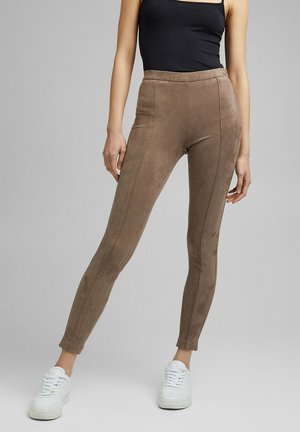 Femme portant un legging en suède marron taille haute et moulant, un haut sans manches noir et des baskets blanches, se tenant devant un fond uni.