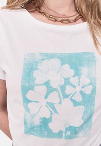 Camiseta blanca de algodón con un estampado floral en turquesa que presenta cinco pétalos y hojas, escote redondo y textura suave. Se puede ver un collar de cadena dorada.