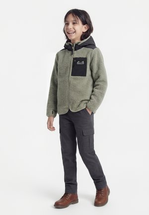 Enfant souriant debout, portant une veste polaire verte avec une poche poitrine noire, un pantalon cargo foncé et des bottes marron à lacets, sur fond blanc.